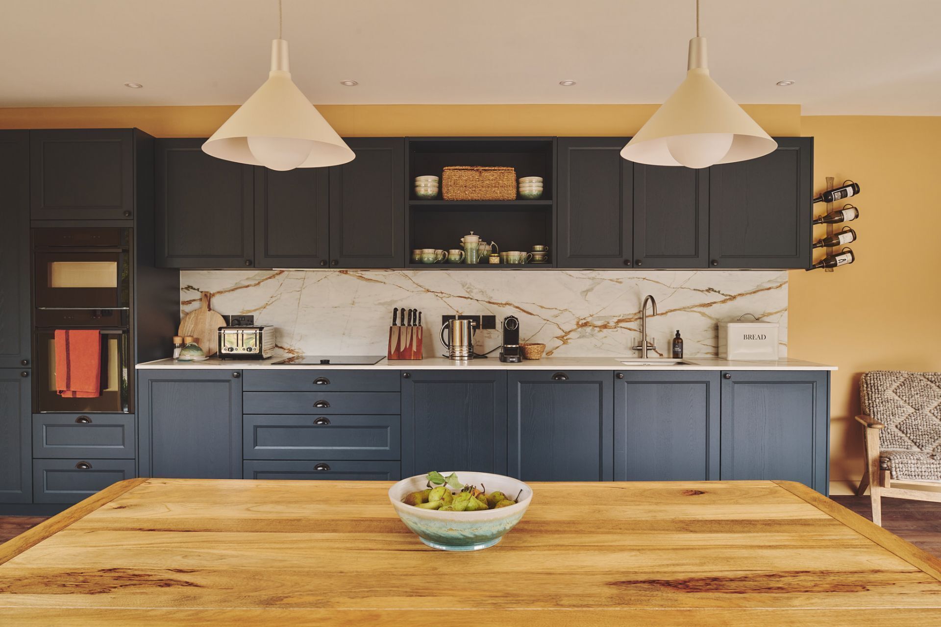 A modern kitchen with dark cabinets, a marble backsplash, and a wooden dining table with a bowl of fruit.