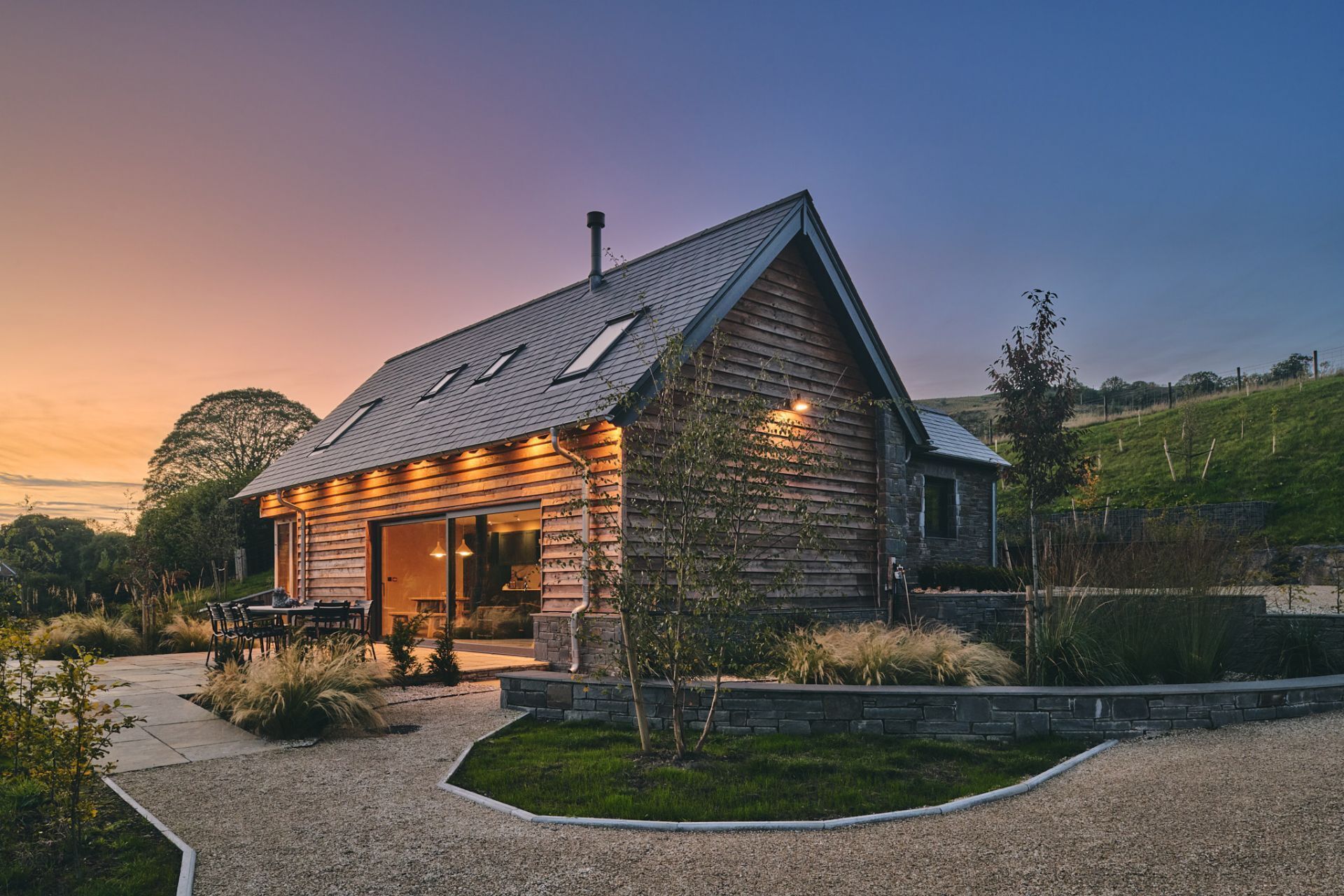 A rustic wooden cabin with a sloped roof, a stone extension, and a gravel driveway.