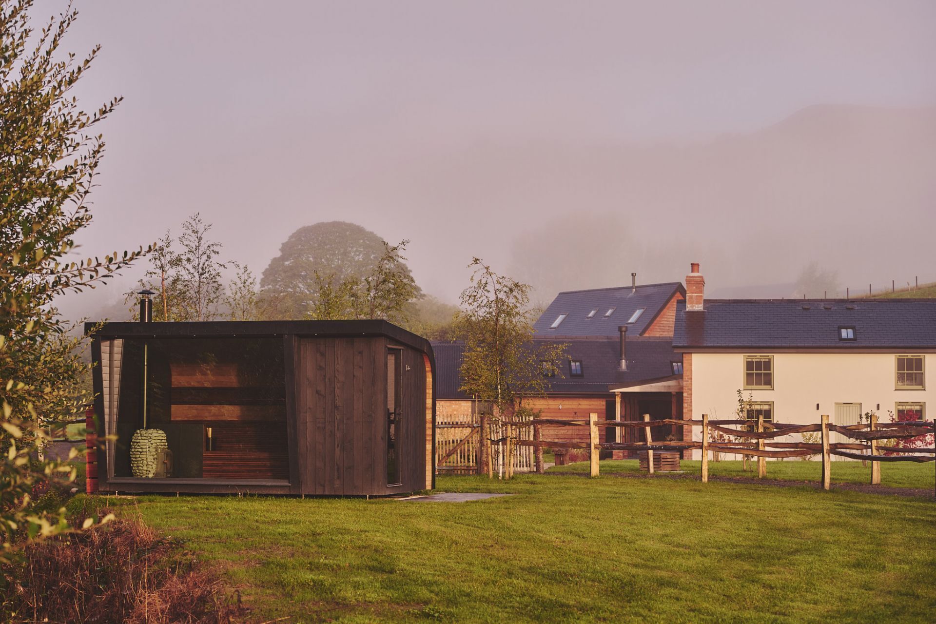 A rustic wooden cabin with a glass door, a fenced yard, and a house with a sloped roof in the background.