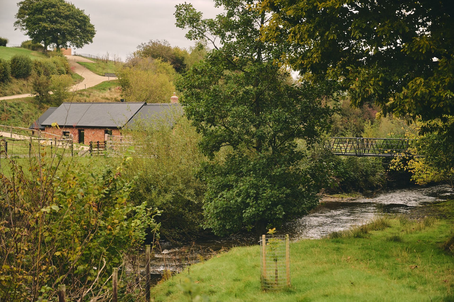 A rural landscape with a river, a wooden bridge, and a building with a sloped roof.