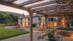 Une terrasse couverte avec une table et des chaises, une maison blanche avec de nombreuses fenêtres et un jardin avec un chemin de gravier.