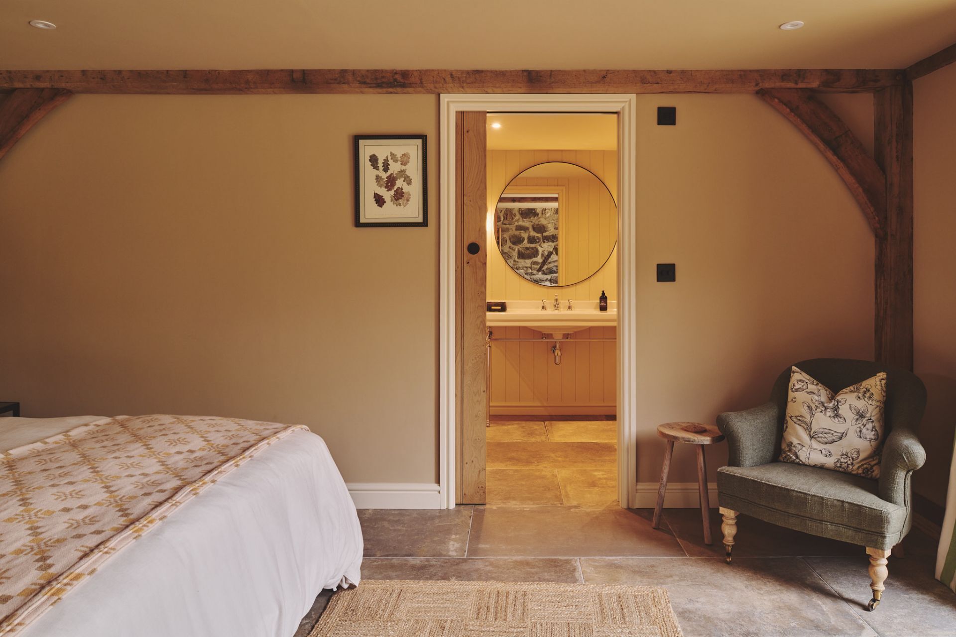 Un dormitorio con una cama, una silla, un taburete pequeño, un espejo y un baño visible a través de una puerta abierta.