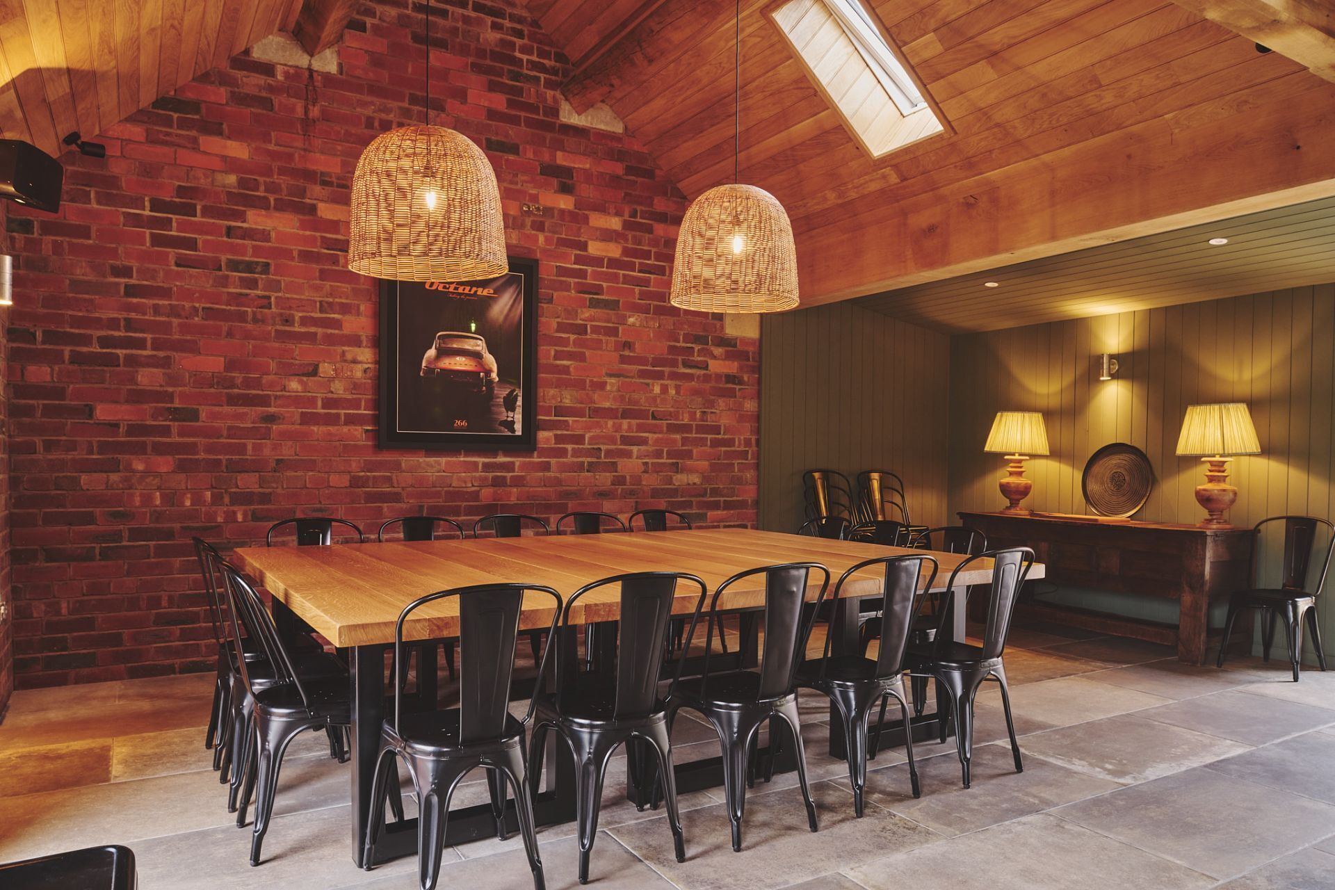 A dining area with a brick wall, wooden ceiling, and a large table with chairs. There are two hanging lamps and a framed picture on the wall.