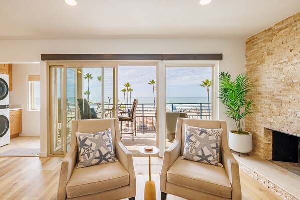 A modern living room with two beige armchairs, a coffee table, and a large sliding glass door leading to a balcony with a view of the sea.