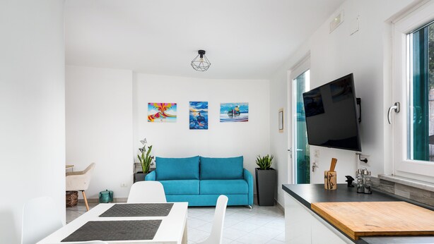 Et moderne køkken med en blå sofa, hvide stole og et spisebord. Der er to potteplanter, et tv ophængt på væggen og billedkunst på væggen.