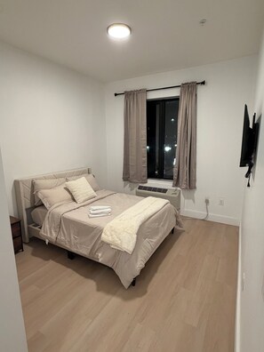 Ein Schlafzimmer mit Bett, einem Fenster mit Vorhängen, einem an der Wand befestigten Flachbildfernseher und einer Deckenleuchte.