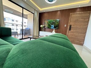Una sala de estar moderna con sofás tapizados en verde, un televisor de pantalla plana montado en una pared de madera, una mesita con un florero y un ventanal corredizo que da a un balcón exterior.