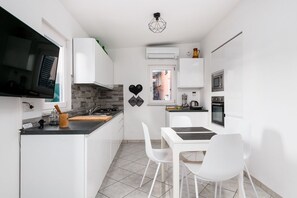 Une cuisine compacte avec des meubles blancs, un plan de travail noir, un coin repas avec des chaises blanches et une petite fenêtre.