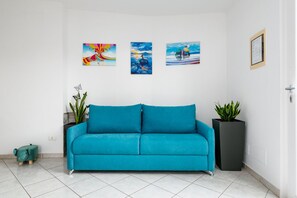 Un sofá color turquesa en una habitación con paredes blancas, tres obras de arte enmarcadas y dos plantas en macetas.