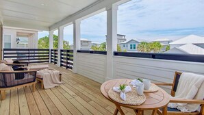 A covered outdoor deck with wooden flooring, a round table with a woven placemat, and two cushioned chairs. - Vibe Out: Private beach steps away, Heated pool, Golfcart, Game room, Bunk rooms (Miramar Beach)