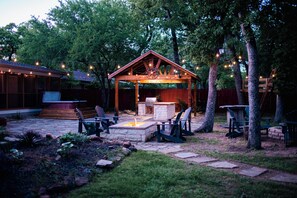 A covered outdoor patio with a stone fire pit, seating, and string lights. - Spacious luxury home in Irving- perfect for groups, families, and Texas getaways (Irving)