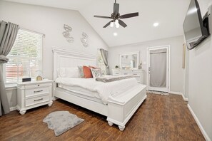 A bedroom with a white bed, a nightstand, a dresser, a ceiling fan, a TV mounted on the wall, and a door.