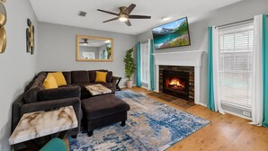 A living room with a sofa, ottoman, coffee table, fireplace, TV, ceiling fan, and a large mirror. - Modern 5BR/2.5BA w/Private Pool  Sleeps 16 ppl (Allen)