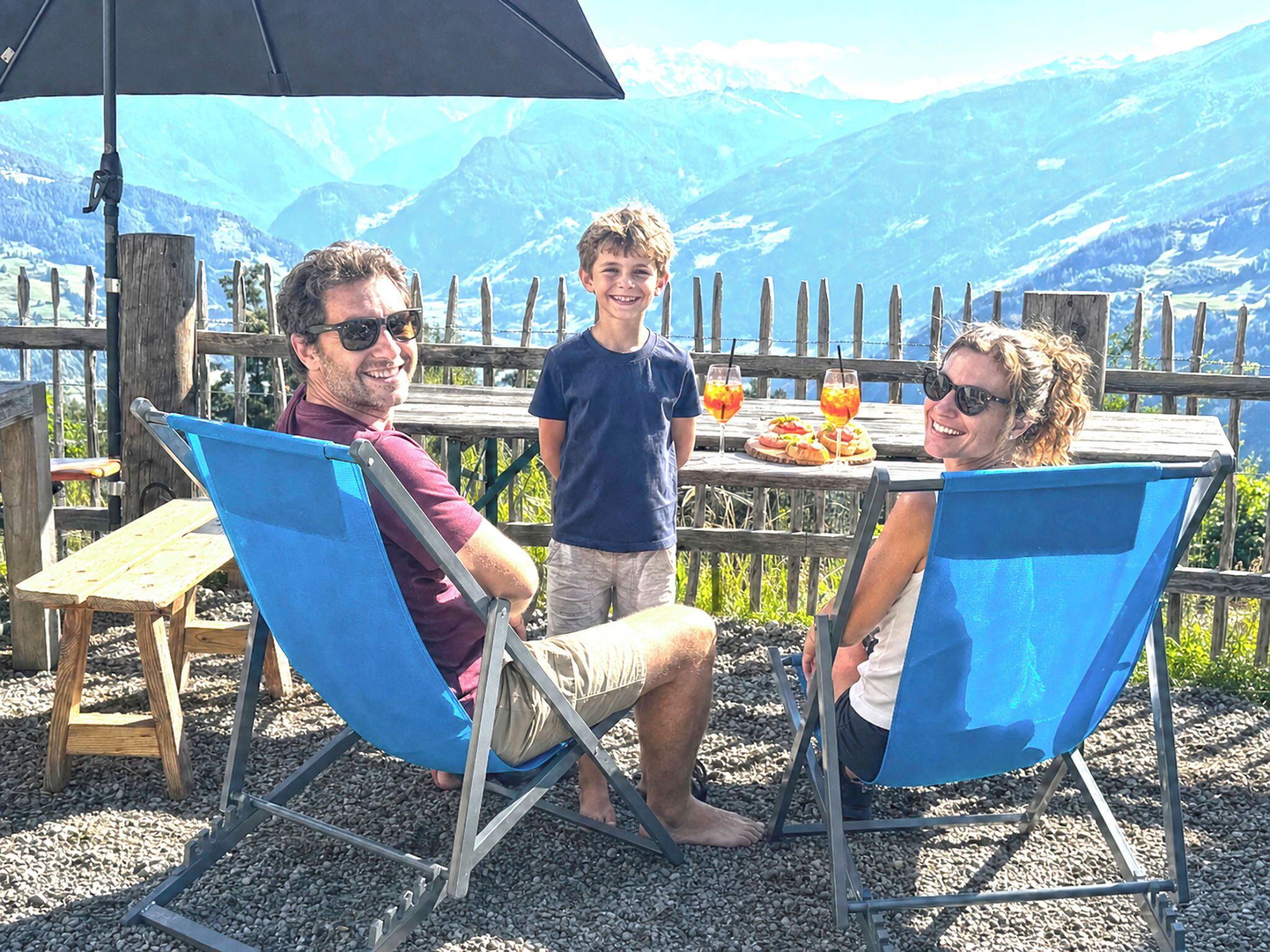 Trois personnes sont assises à une table à l’extérieur, avec une vue sur la montagne, une clôture en bois et une table garnie de nourriture et de boissons.