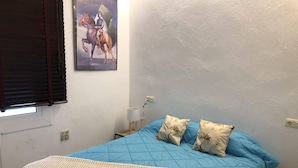 A bedroom with a blue bedspread, two pillows, a white textured blanket, a painting of a horse on the wall, a window with blinds, and a bedside lamp. - Apartment in Roses on the seafront.MONTEMAR (Roses)
