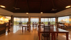 A spacious dining area with large windows offering ocean views, wooden tables and chairs, and ceiling fans. - Balangan Surf Resort (Jimbaran)