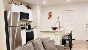 Una cocina moderna con un refrigerador de acero inoxidable, microondas y horno, un sofá gris con varios cojines, y una pequeña isla con un lavamanos y un cuenco decorativo.