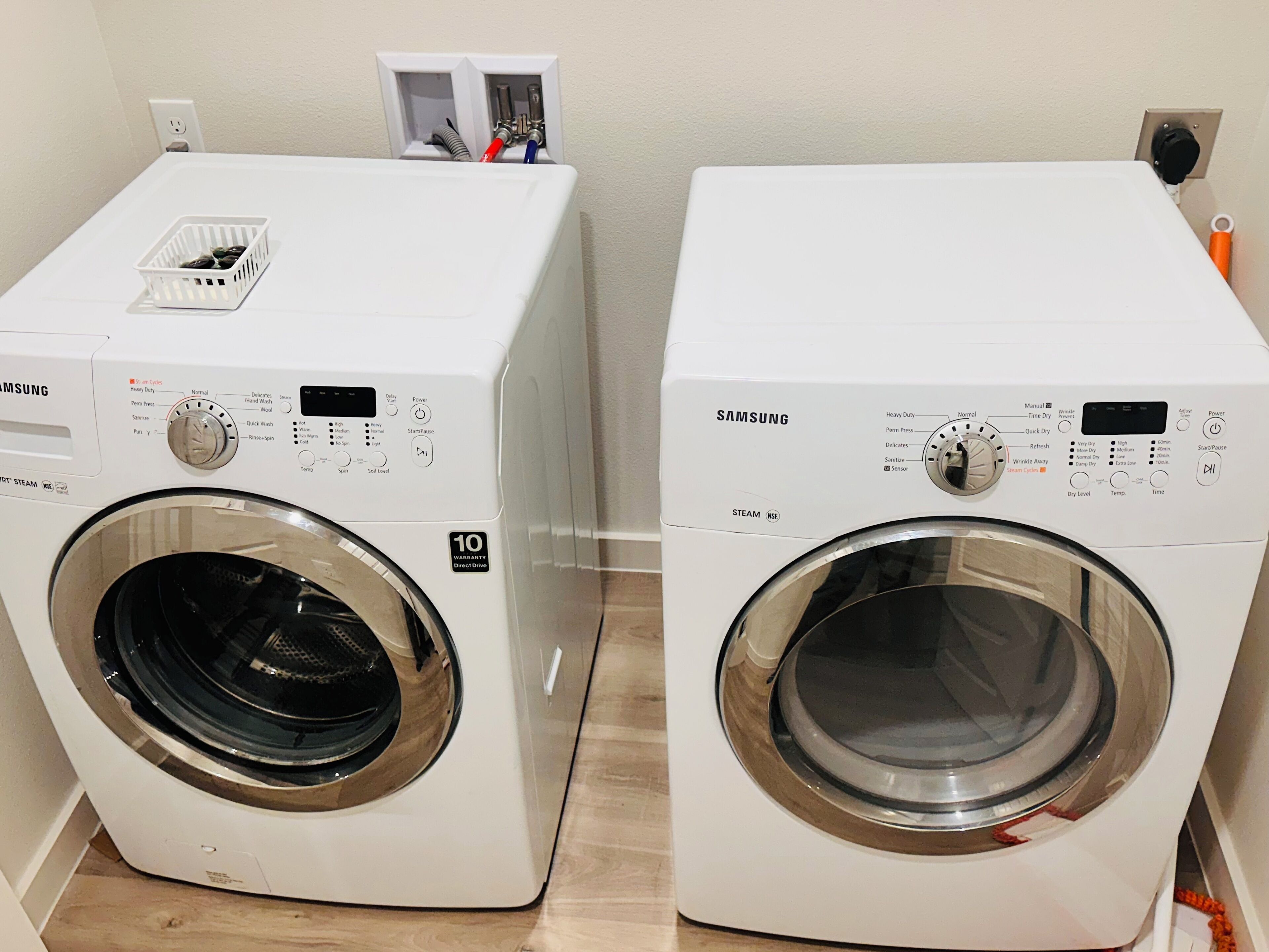Two white Samsung front-loading washing machines side by side with a control panel, a basket, and a power outlet in the background.