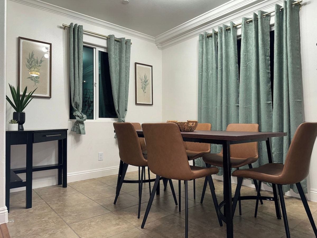 A dining area with a table and chairs, a small side table, and curtains on the windows.