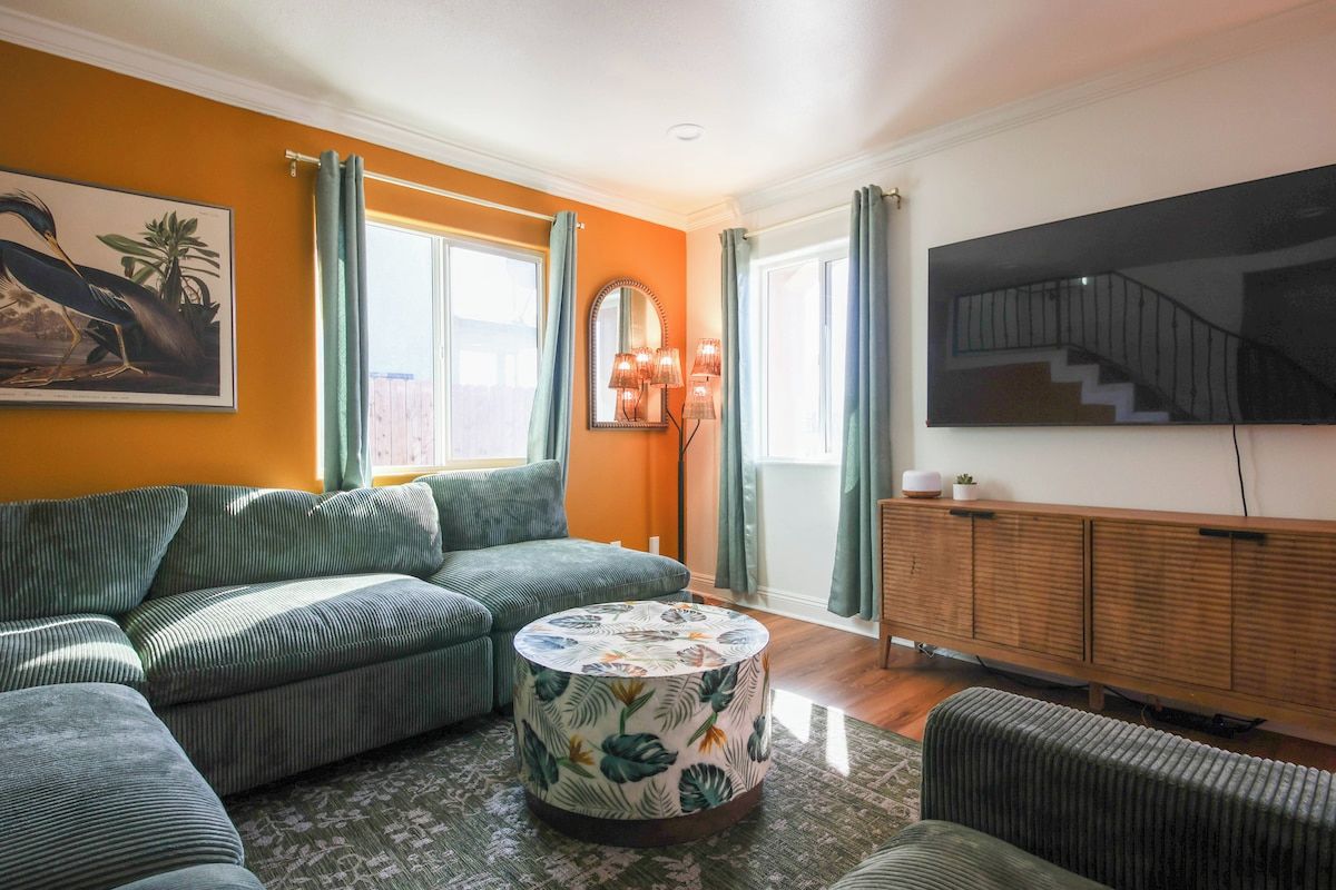 Ein Wohnzimmer mit einer Eckcouch, einem runden Couchtisch mit Blumenmuster, einer Holzkommode und einem großen Flachbildfernseher an der Wand.