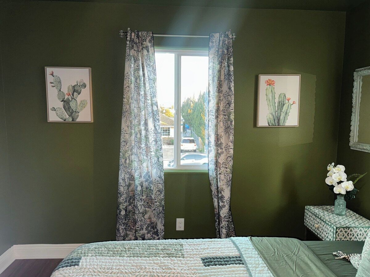 A bedroom with a bed, curtains, a window, a cactus painting, and a vase with flowers.