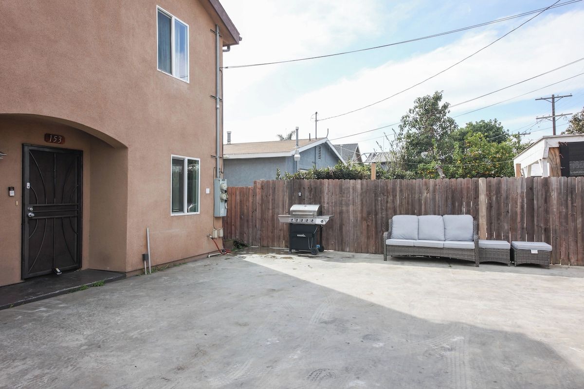 A residential area with a concrete driveway, a wooden fence, a barbecue grill, and a grey sofa.