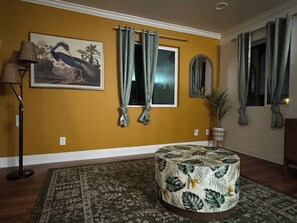 Ein Zimmer mit einer gelben Wand, einem großen Gemälde, einem runden Hocker mit tropischem Muster, einer Stehlampe und einem Fenster mit Vorhängen.