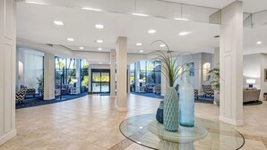 Eine geräumige Lobby mit einem Glastisch, einer Sitzecke mit gemusterten Kissen und großen Fenstern für natürliches Licht.