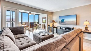 Ein geräumiges Wohnzimmer mit einer großen, grauen Eckcouch, einem Essbereich mit Holzsesseln, einem Fernseher und einem Balkon mit Blick.