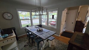 Un coin repas avec une table en bois, des chaises et une horloge au mur. Il y a des fenêtres donnant sur l’extérieur.
