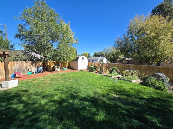 Un patio trasero con cerca de madera, un cobertizo pequeño, una zona de juegos con un tobogán y un jardín con camas elevadas.