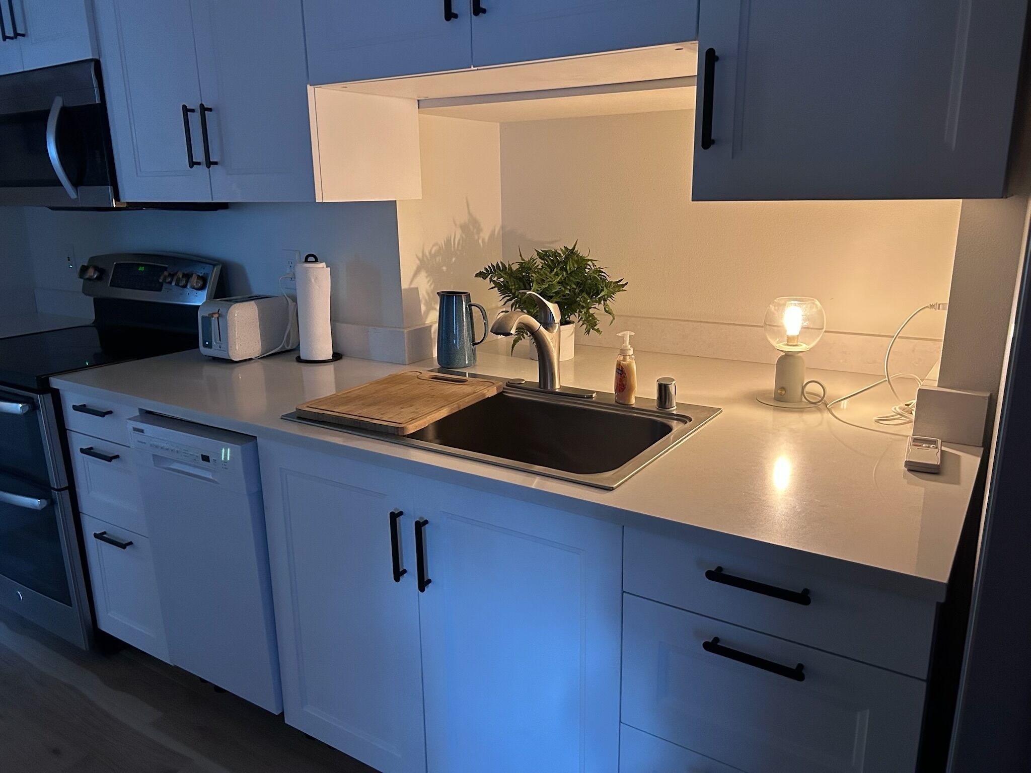 A modern kitchen with white cabinets, a stainless steel sink, a wooden cutting board, a kettle, a toaster, a plant, and a light fixture.