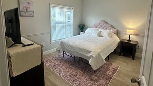 Un dormitorio con cama, mesita de noche, lámpara, ventilador de techo, ventana con persianas, un cuadro y televisor sobre un soporte.