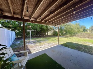 En udendørs overdækket terrasse med træloft, en hvid bænk og et område med grønt kunstgræs.