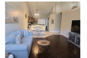 A living room with a sofa, a coffee table, and a TV stand. There is a dining area with a table and chairs in the background. - Adv. Coast - 2BR/2BH Near Springs, Beach, Fenced (Spring Hill)