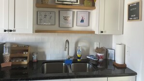Cucina con mobili bianchi, lavello in acciaio inossidabile, ripiano in legno con opere d'arte incorniciate, una pianta in vaso e un cartello di benvenuto.