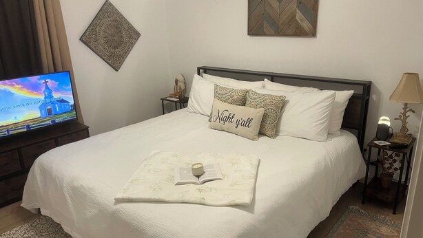 Un letto rifatto con cura, un cuscino con la scritta “Night y'all”, una lampada da comodino, un tavolino con un oggetto decorativo e un televisore a parete che mostra un'immagine panoramica.