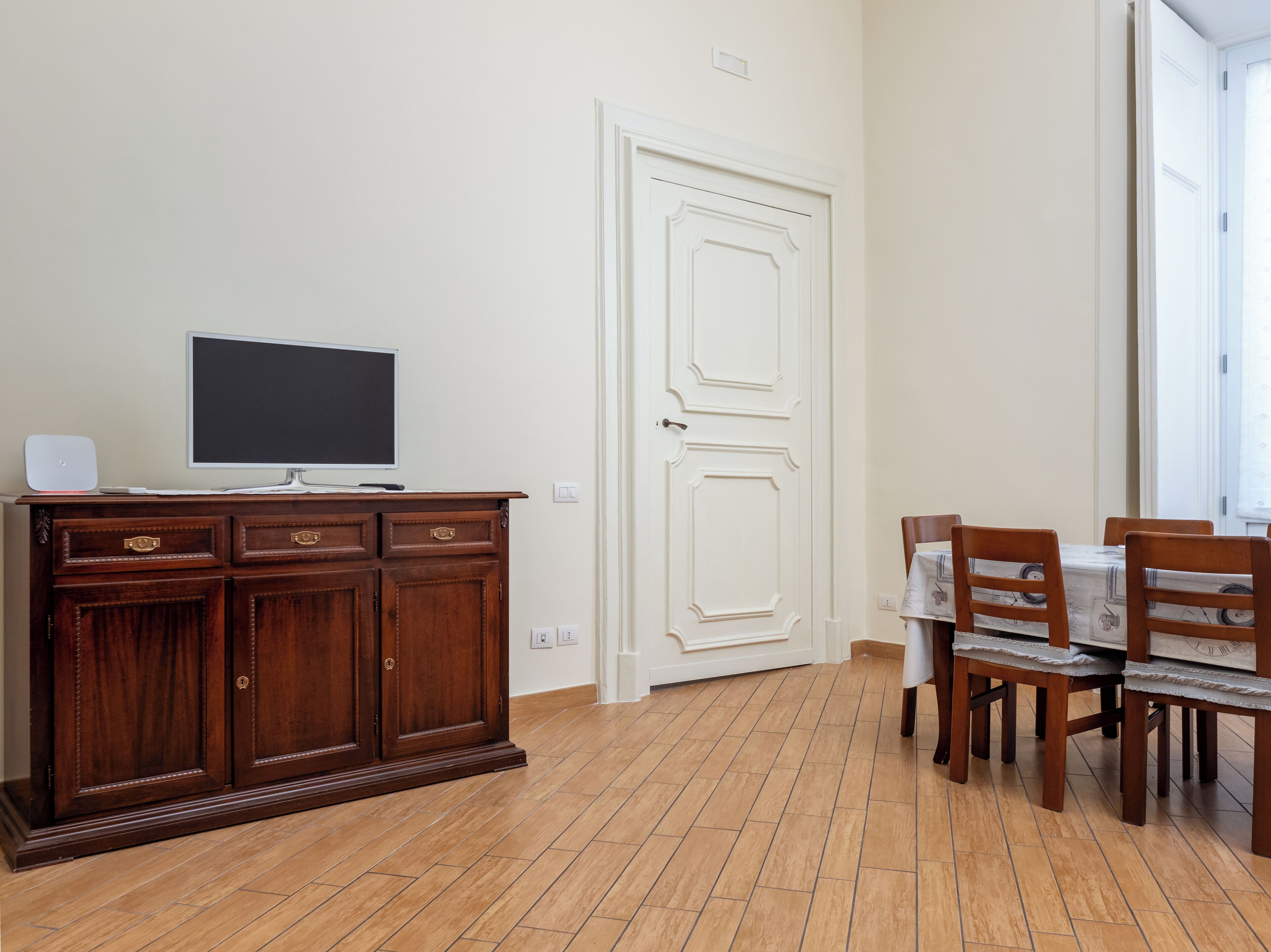 A room with a wooden cabinet, a flat-screen TV, a dining table with chairs, and a white door.