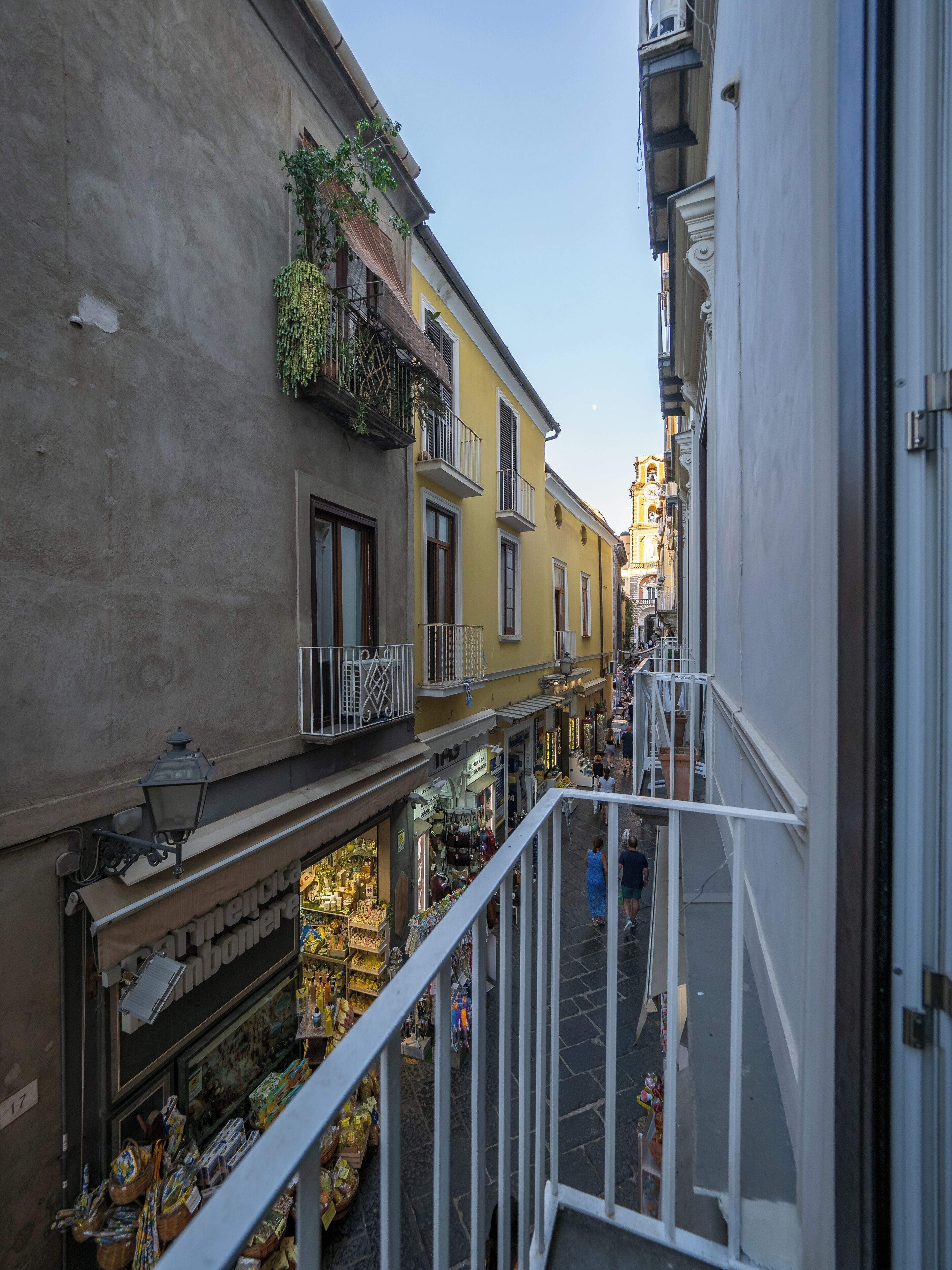 A narrow street with buildings on either side, balconies with plants, and a shop with various items on display.