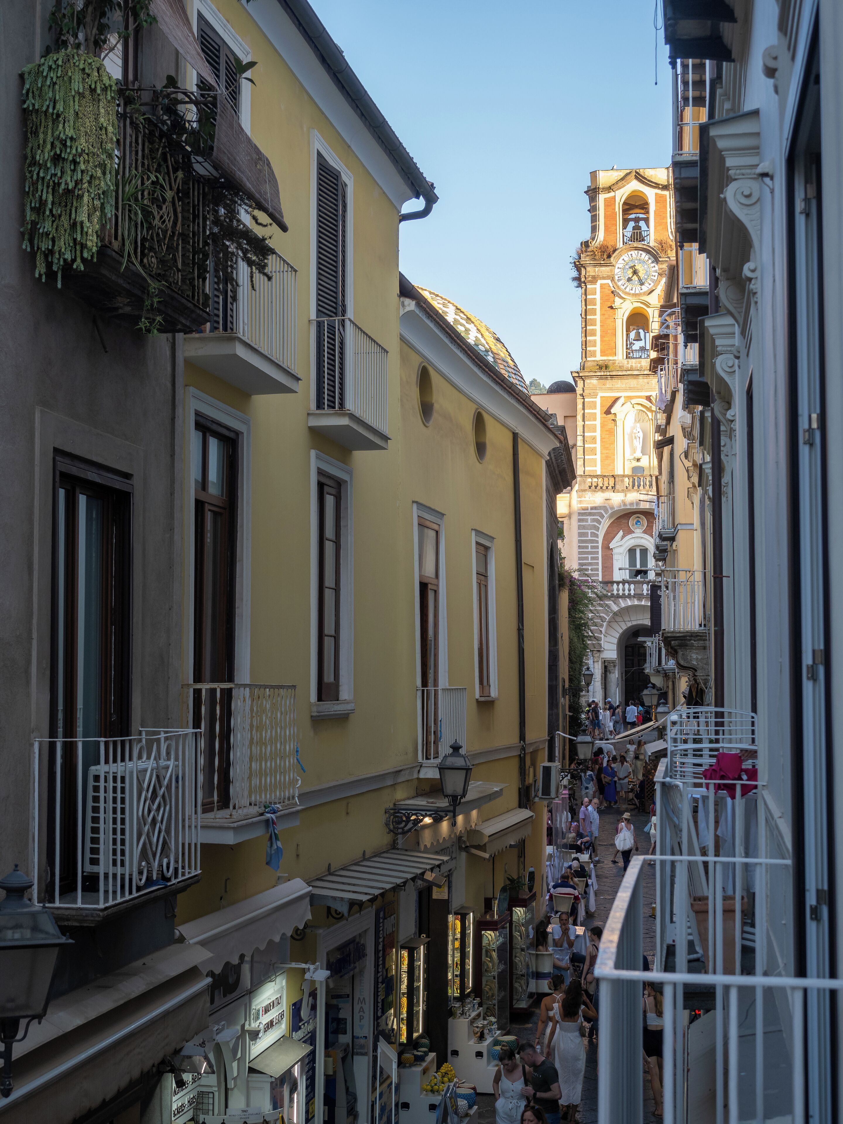 A narrow street with yellow buildings, a clock tower, and balconies with plants.