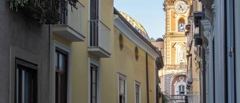 A narrow street with yellow buildings, a clock tower, and balconies with plants.