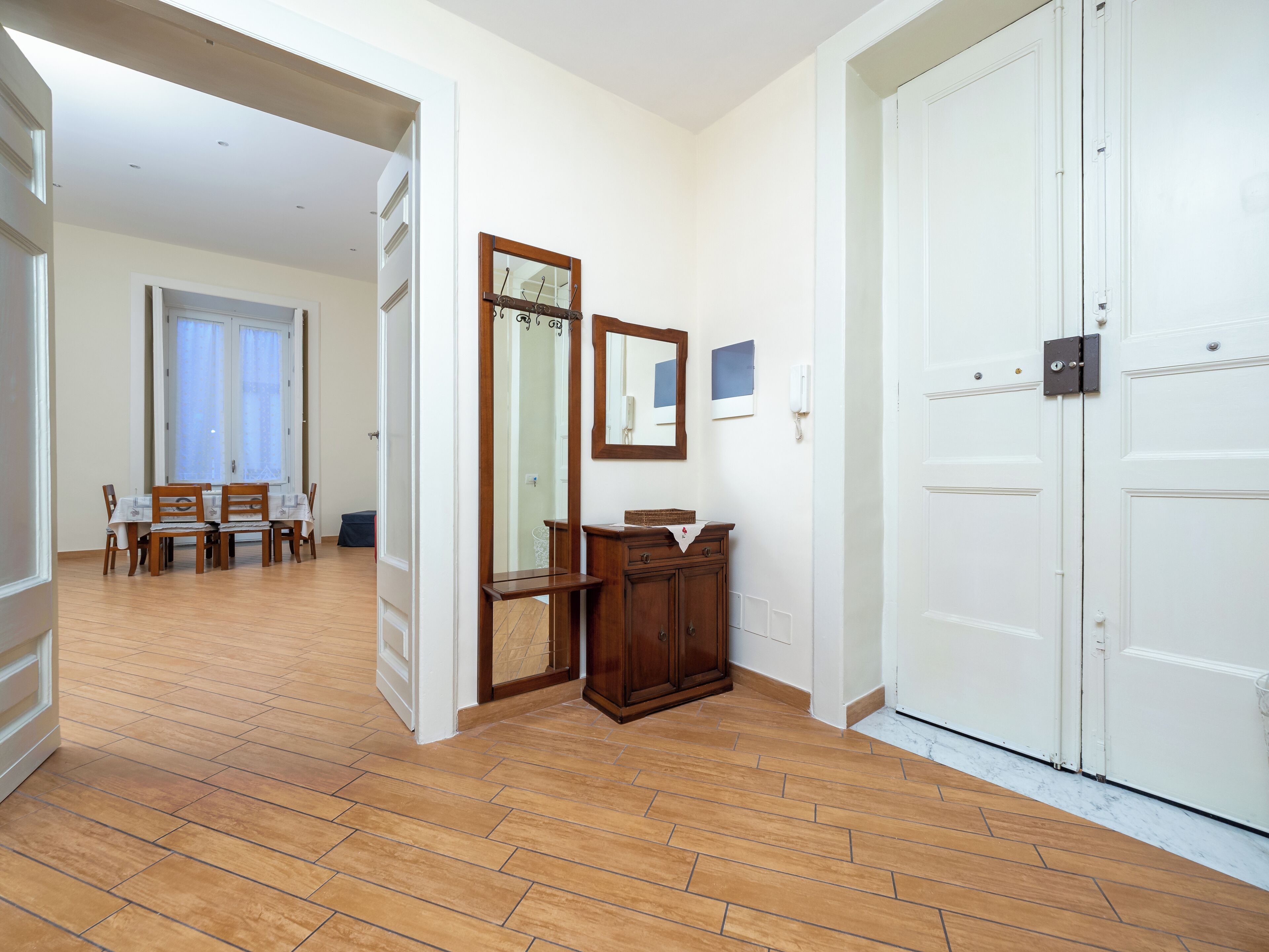 A hallway with wooden flooring, a dining area with a table and chairs, and a white door with a lock.