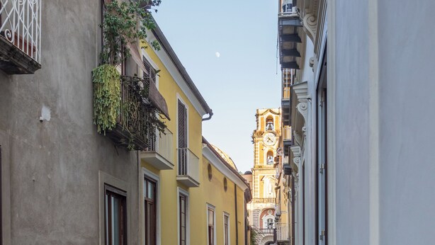 A narrow street with shops on either side, a clock tower in the distance, and a pedestrian walkway. - Maya Home (Sorrento)
