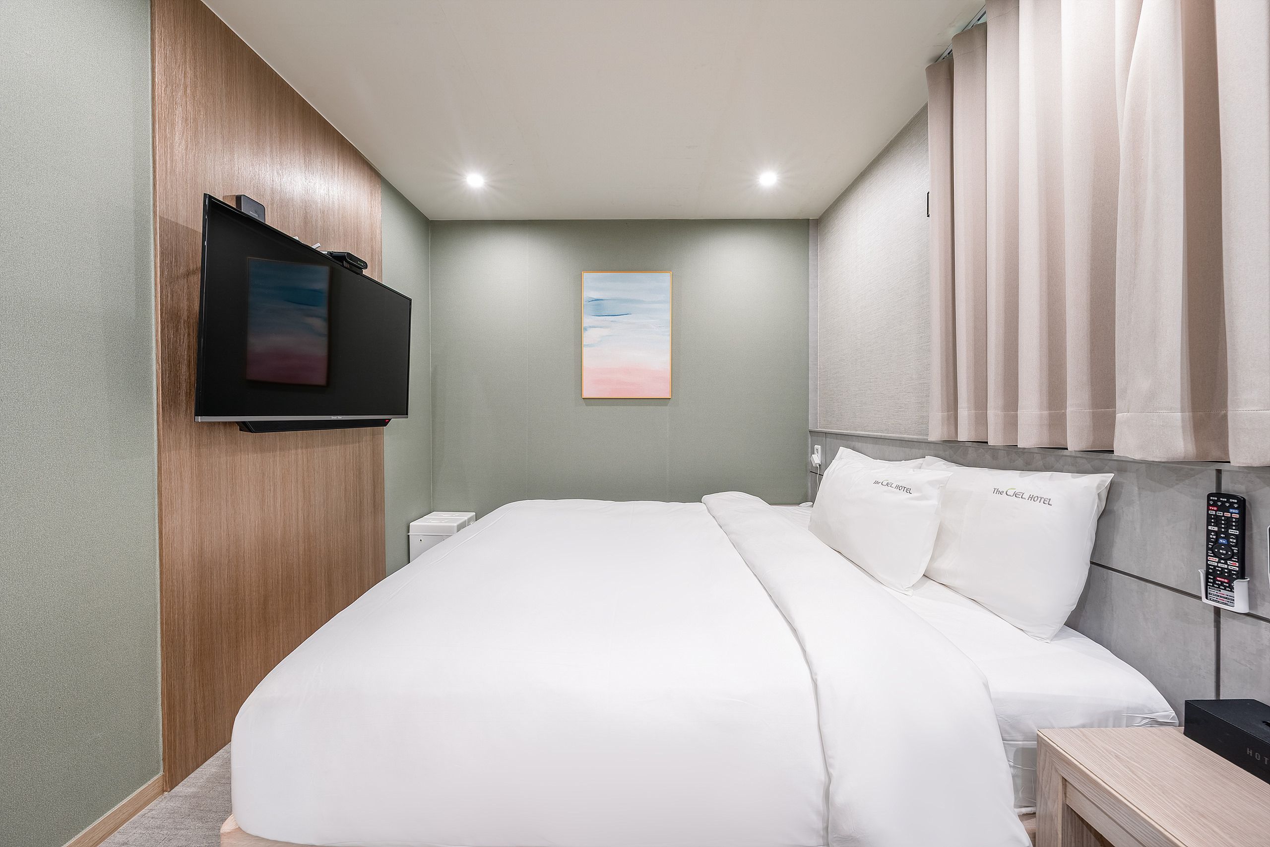 A hotel room with a bed, a TV mounted on the wall, a painting above the bed, and a window with curtains.