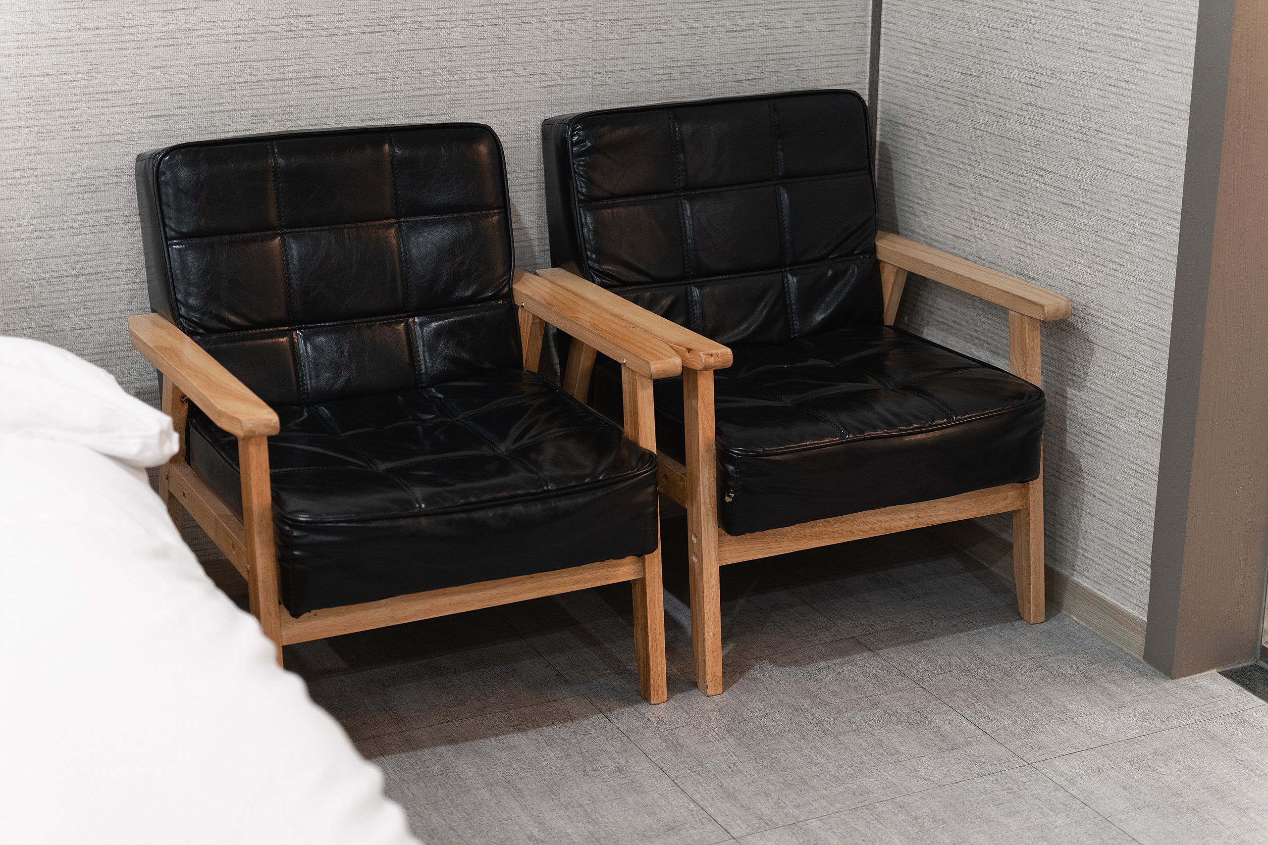 Zwei schwarze Ledersessel mit Holzarmlehnen und -beinen, platziert in einer Ecke des Raumes mit grauem Teppichboden und einer strukturierten Wand in Grau.