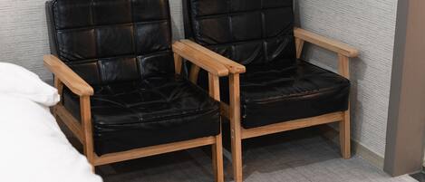Two black leather chairs with wooden armrests and legs, positioned in a corner of a room with grey carpeted flooring and a grey textured wall.