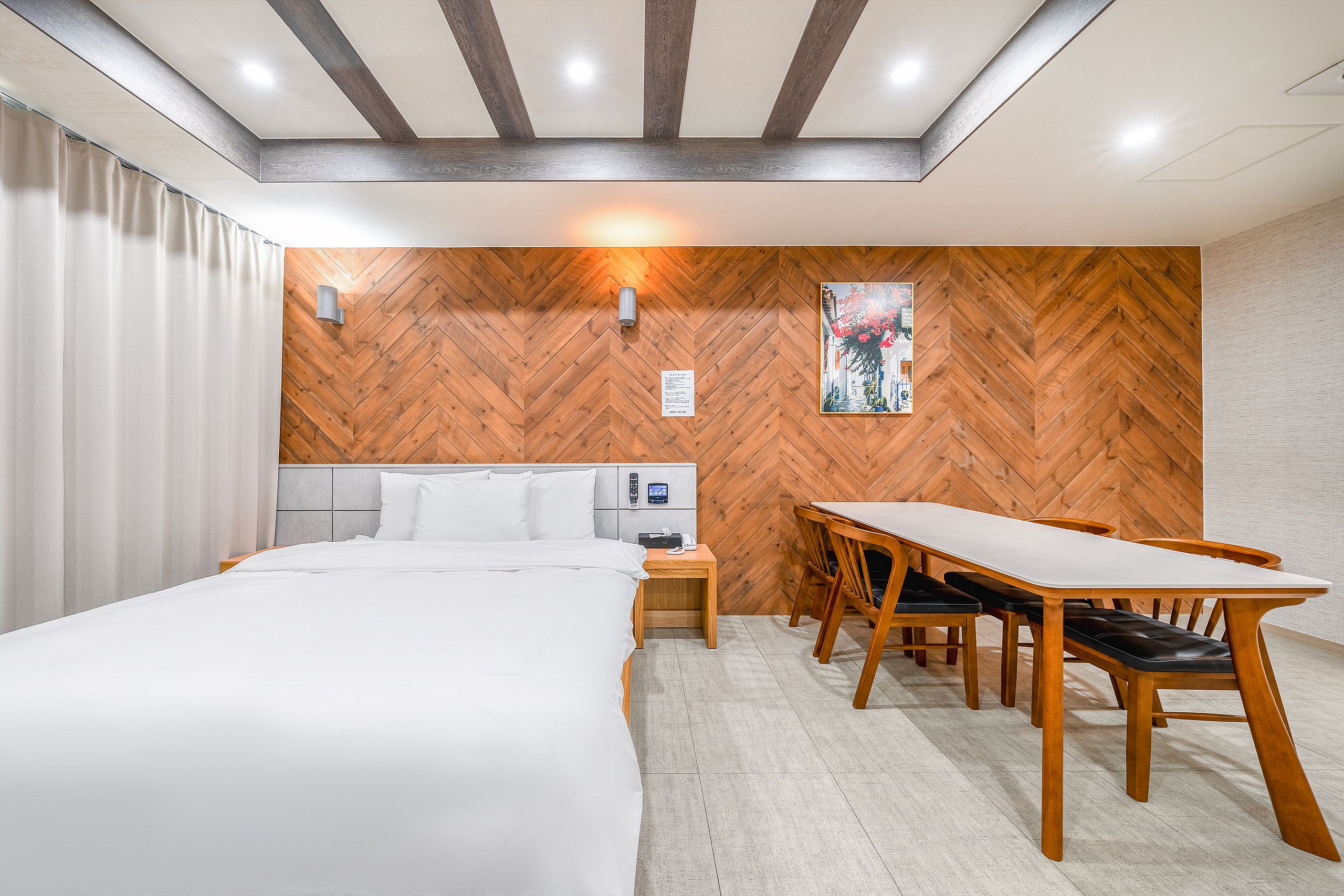 A hotel room with a bed, a desk, and a chair. There is a wooden herringbone wall and a white curtain.