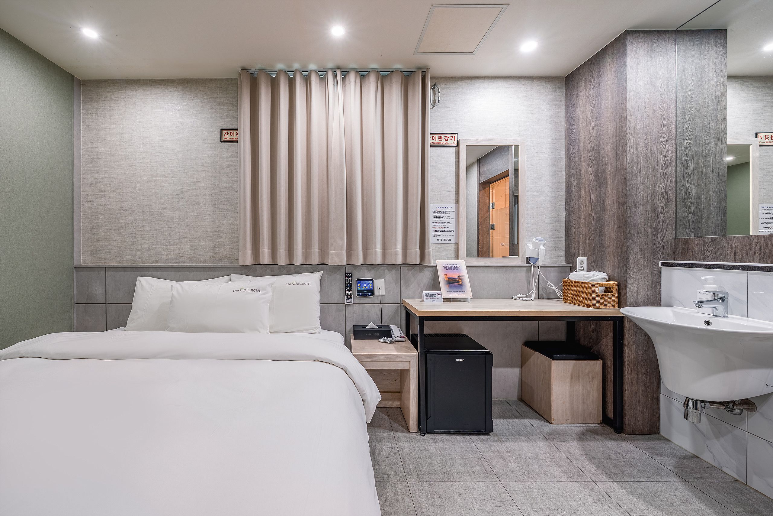 Habitación de hotel con cama, escritorio, un basurero pequeño, espejo y baño con lavamanos e inodoro.