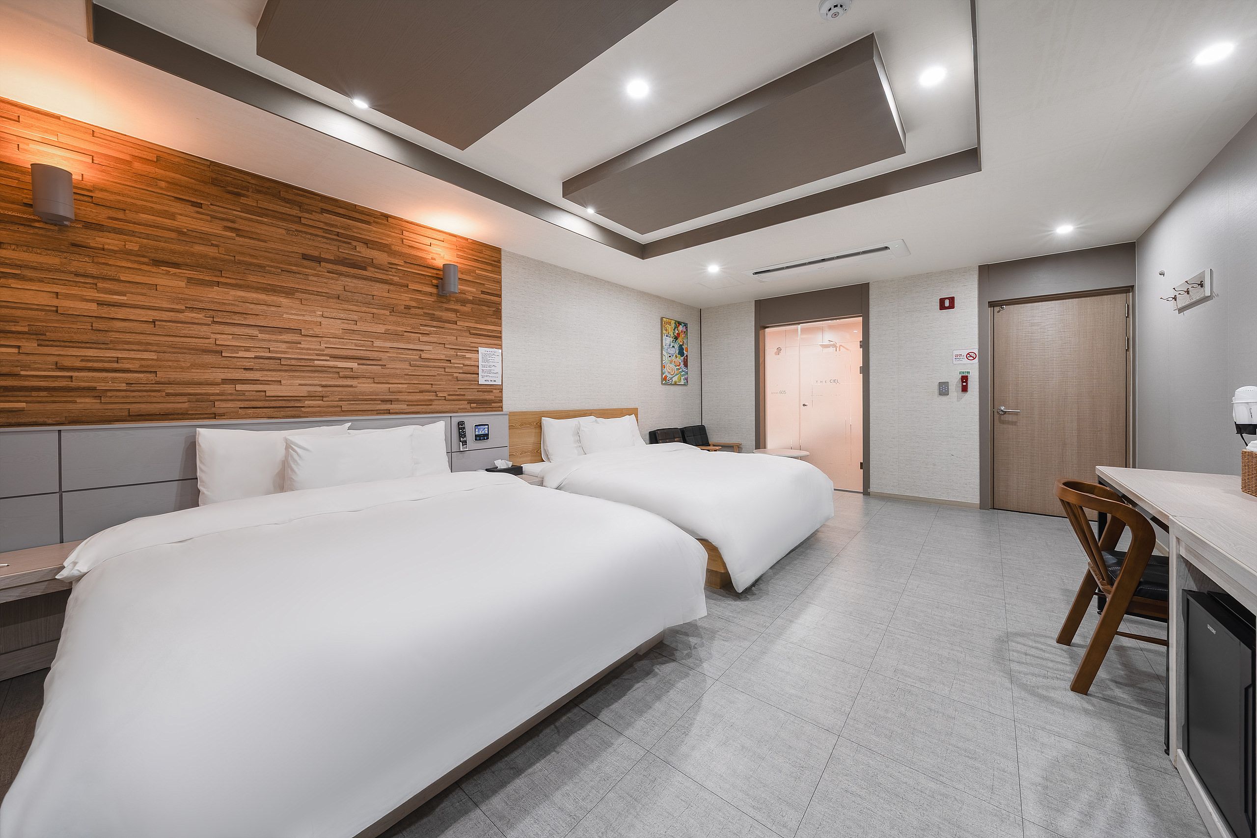 A hotel room with two beds, a wooden accent wall, a small desk, and a bathroom door.