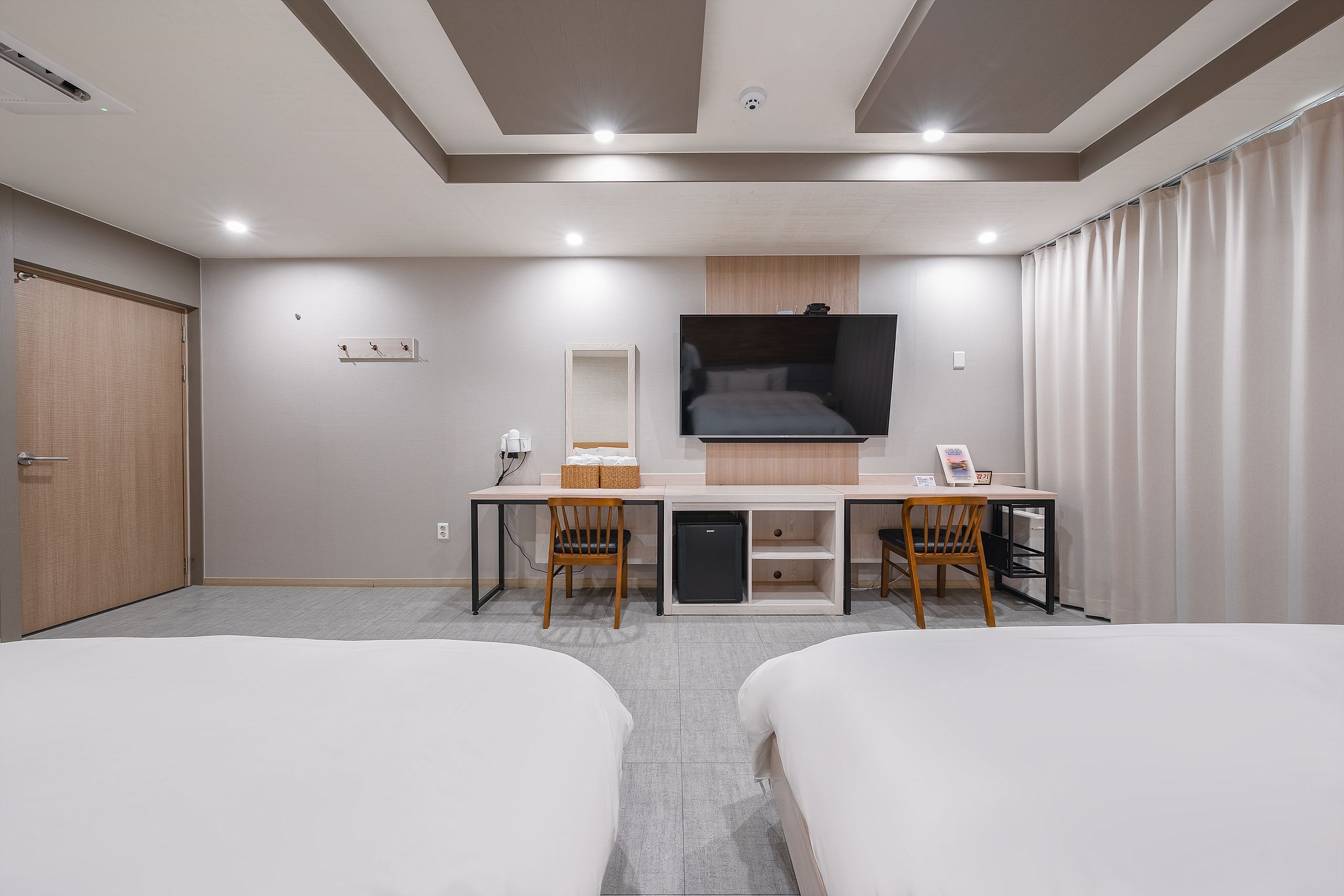 A hotel room with two beds, a desk with a flat-screen TV, a small refrigerator, and a wooden cabinet.